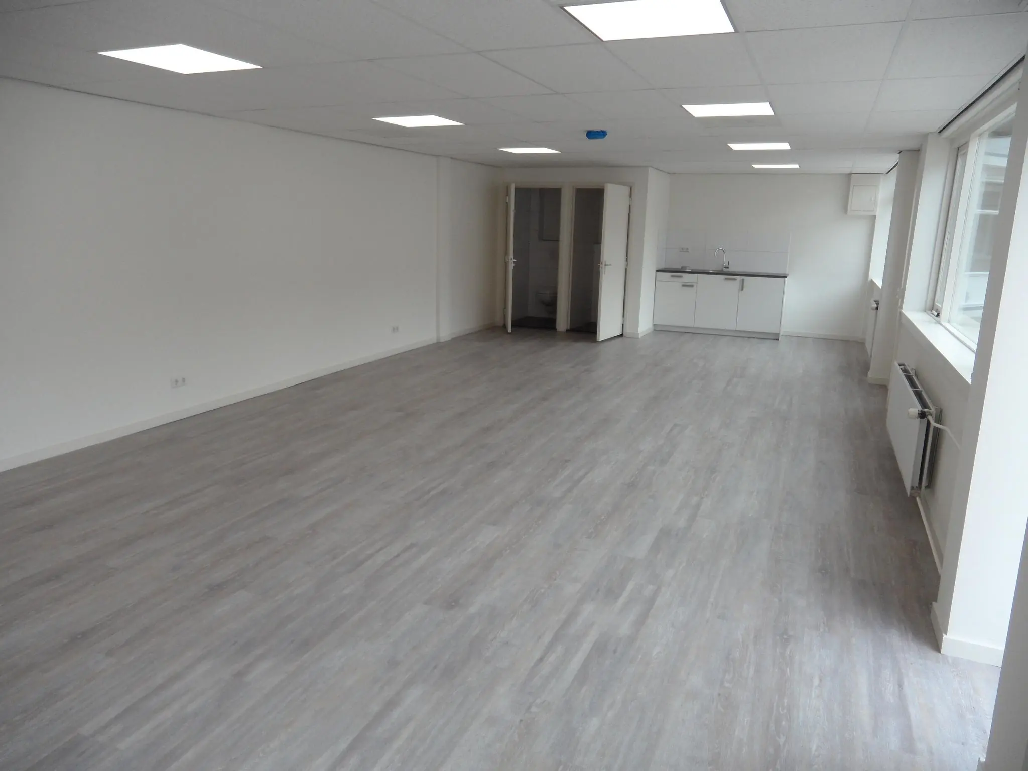 Lichte, lege kantoorruimte aan de Jacques Perkstraat met grijze vloer, systeemplafond en een kleine keuken.